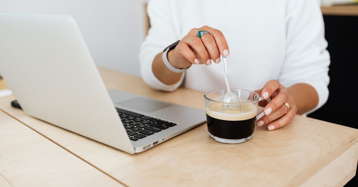 A woman stirs coffee at her desk while working on a laptop in a modern office setting.