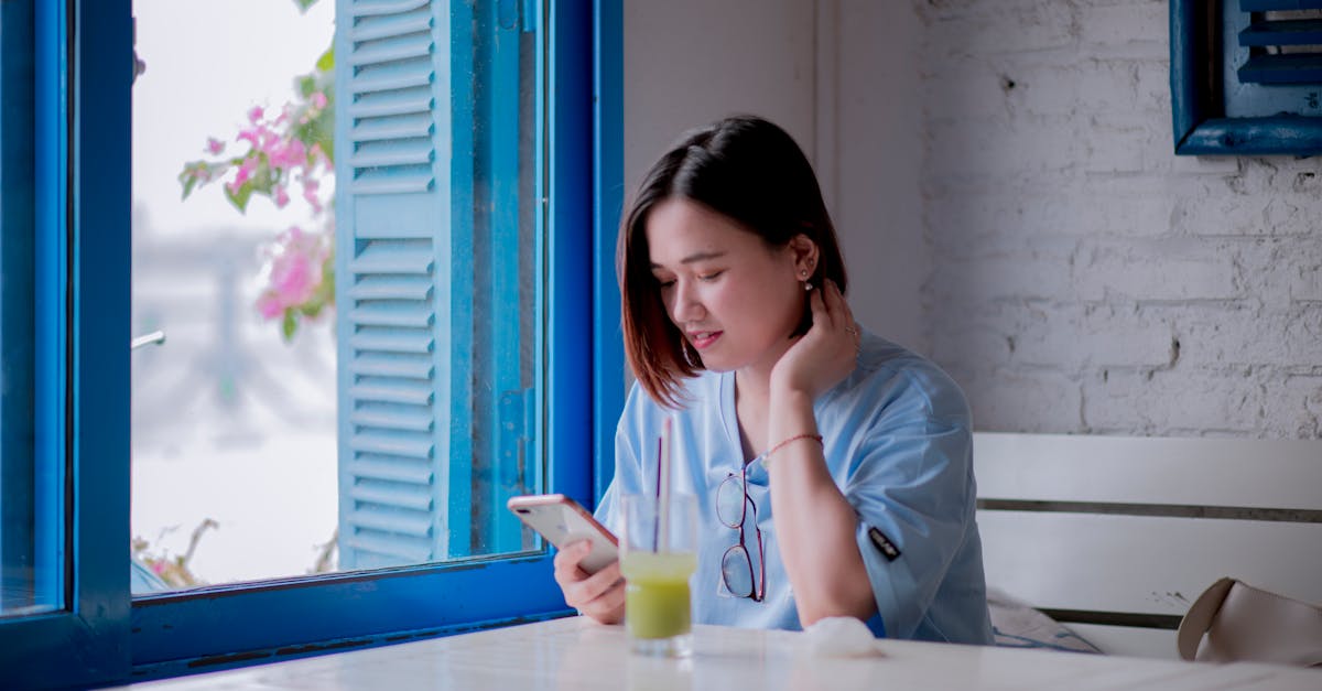 A young woman in a light blue shirt checks her smartphone while enjoying a drink by a window in a quaint cafe.